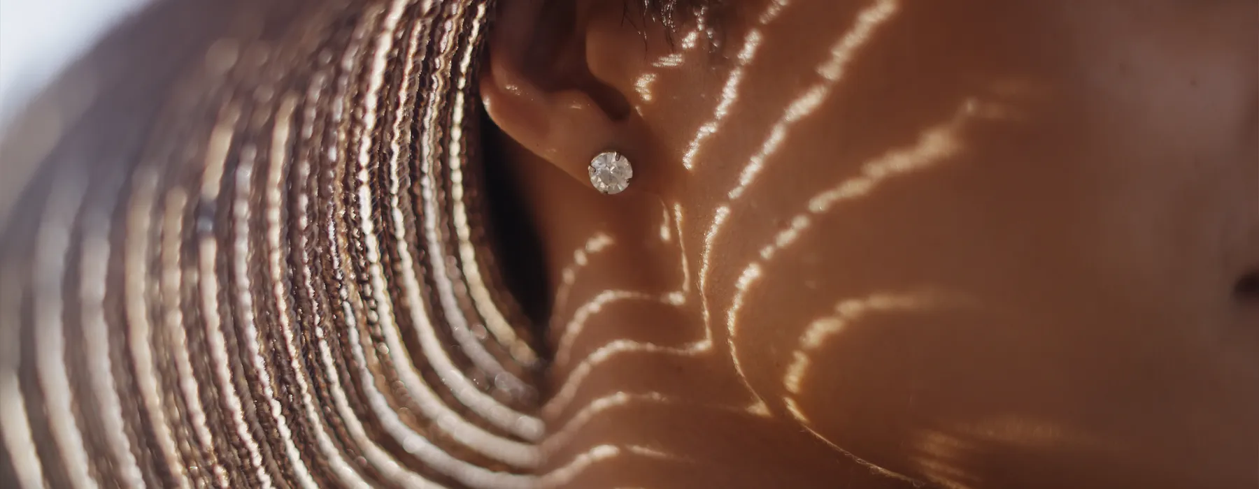Close-up of a woman's ear with a diamond earring, sunlight casting wavy shadows on her skin.