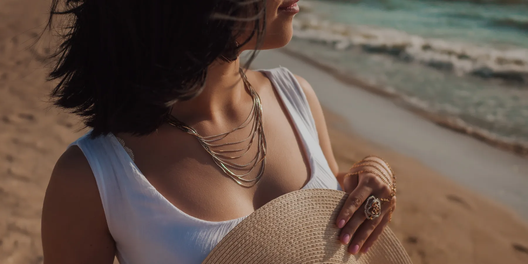 A woman in a white dress holds a straw hat, adorned with layered necklaces and rings, by the beach. Explore Our Jewelry.
