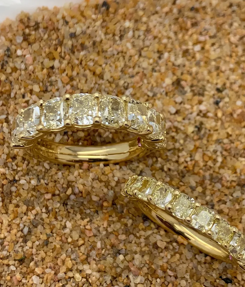 Two gold rings with rectangular diamonds set on a sandy surface.