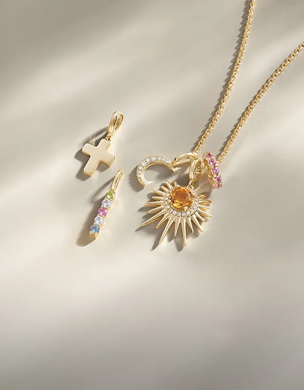 Gold necklace with various charms, including a cross, crescent, and sunburst, on a light background.