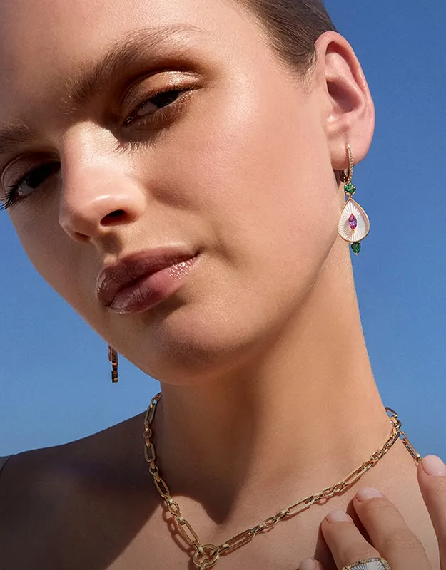 A woman wearing gold earrings with pink and white gemstones, a gold chain necklace, and a ring against a blue sky.