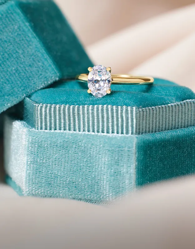 A gold engagement ring with an oval diamond on a teal velvet box.