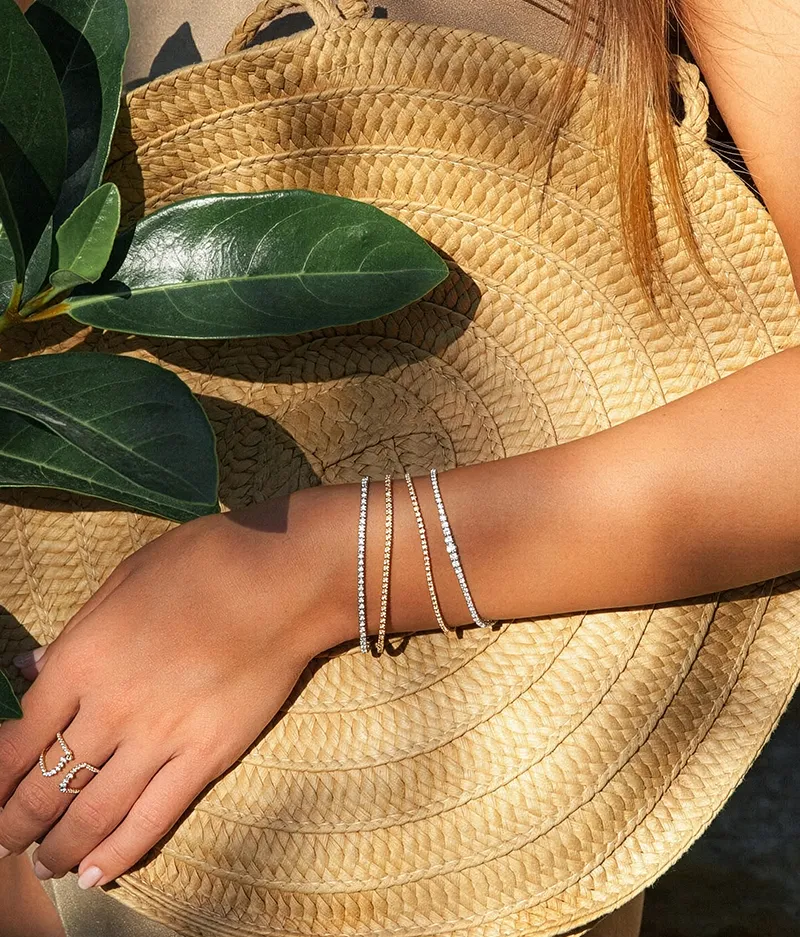 A hand with diamond bracelets and ring holds a woven straw bag, next to green leaves.