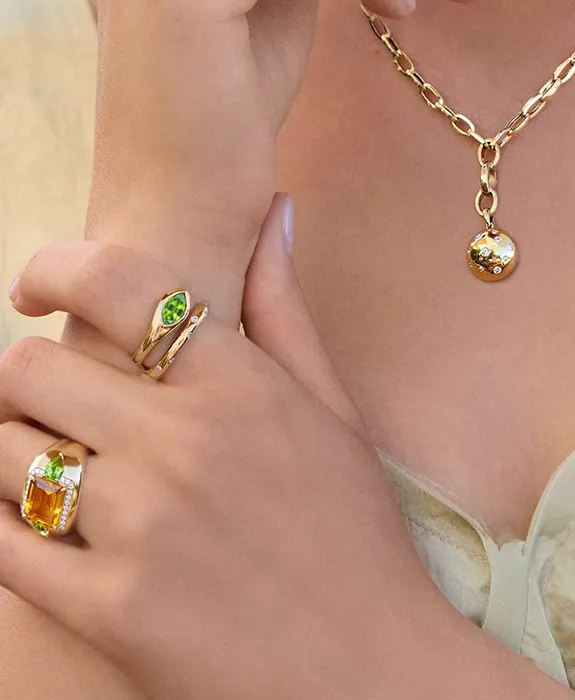 Woman wearing gold necklace and rings with green and orange gemstones.