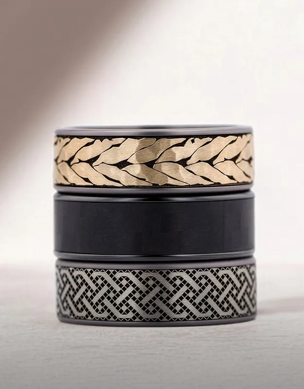Three men's wedding bands stacked: gold with leaf pattern, plain black, and silver with intricate design.