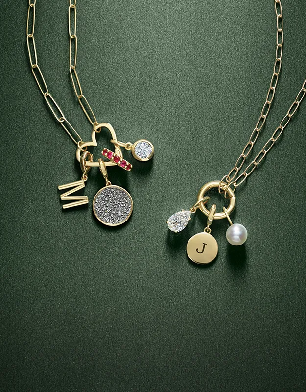 Two gold necklaces with charms, including initials, a pearl, and gemstones, on a green background.