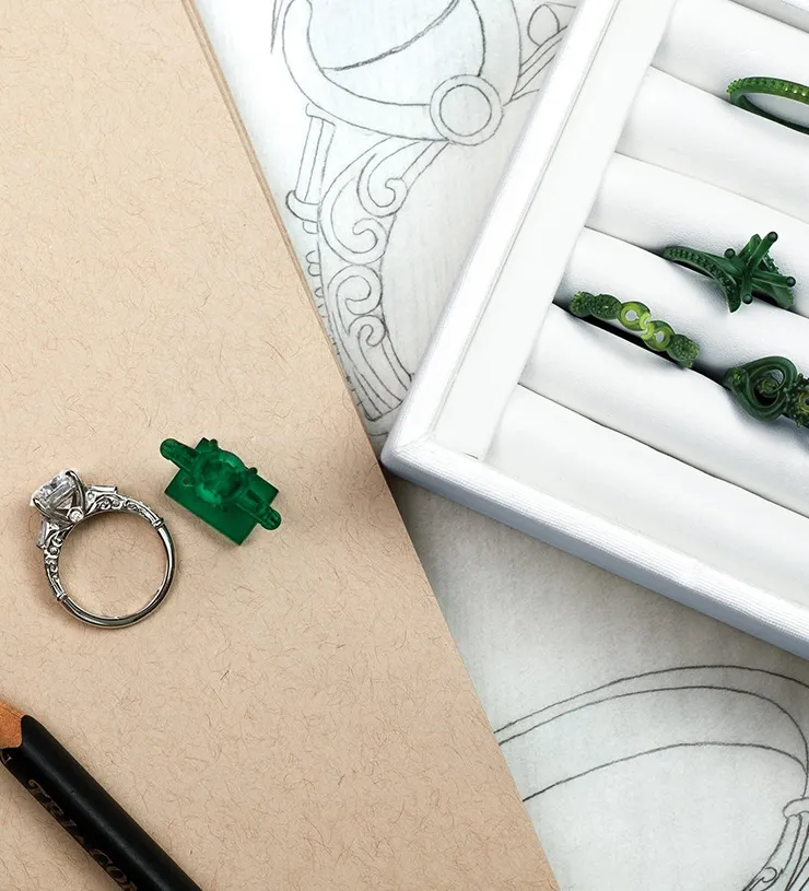 A silver ring with a gemstone, green wax molds, and ring sketches on a table.