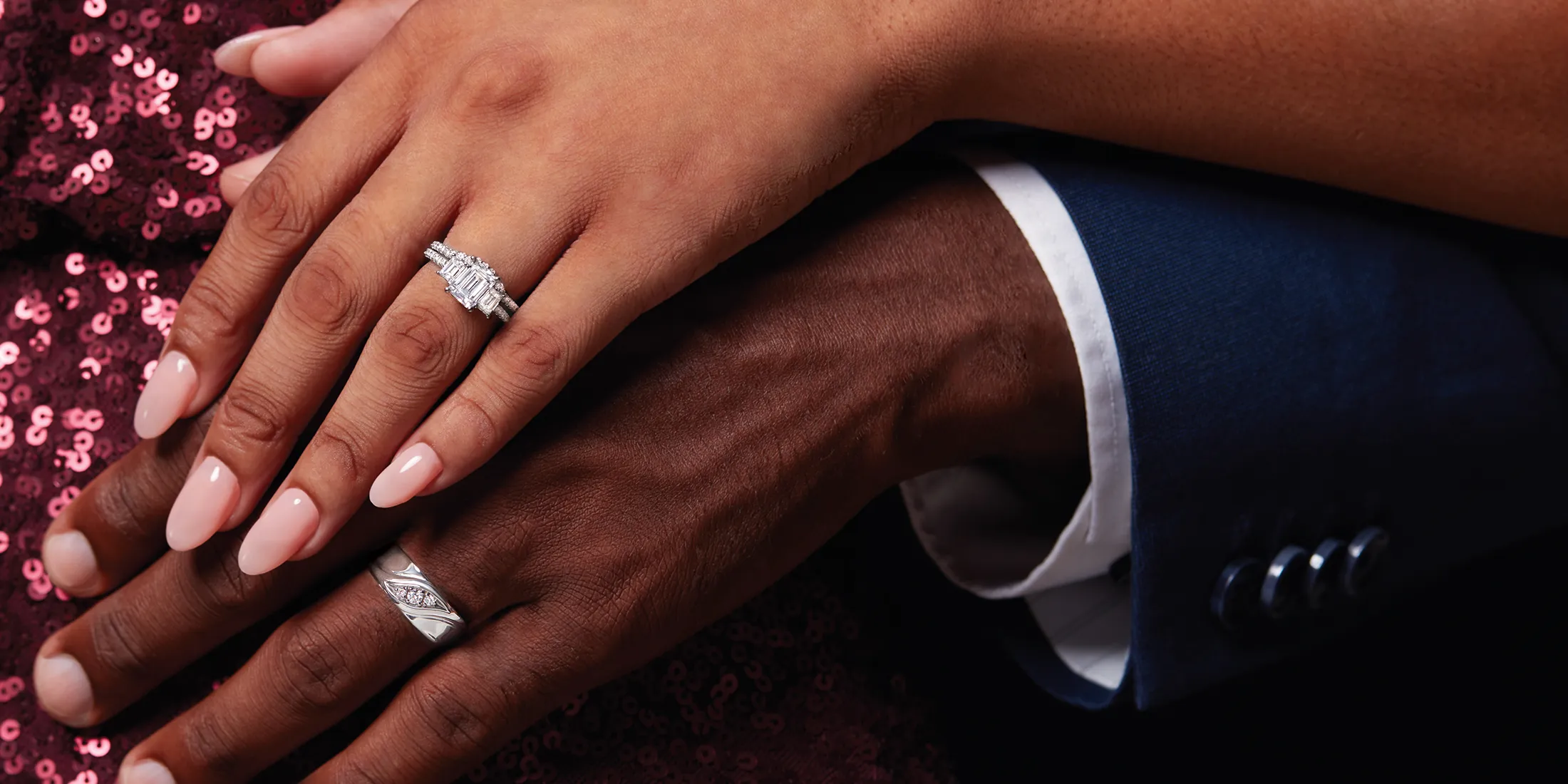 Two hands with wedding rings, one with a diamond ring, resting on a sequined fabric.