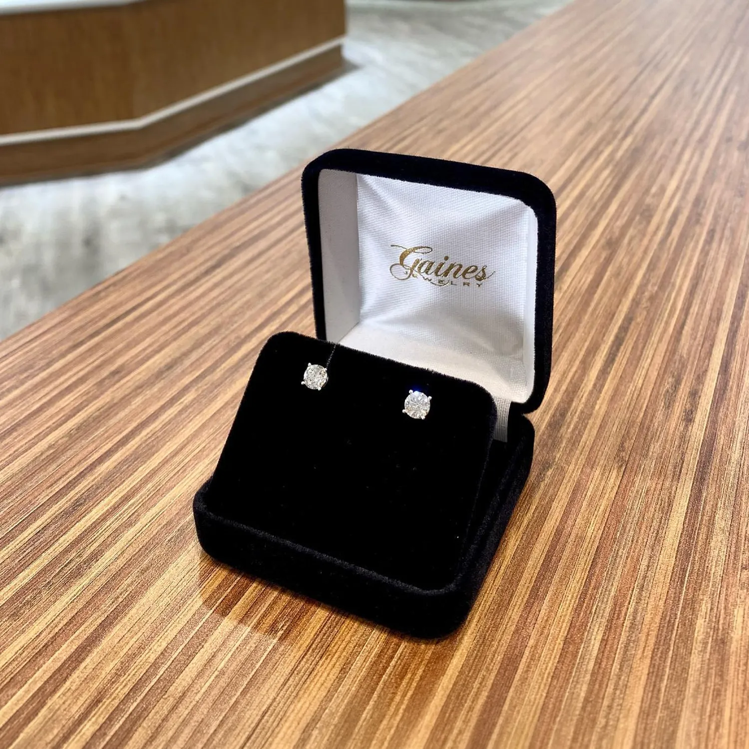 Diamond stud earrings in a black velvet box on a wooden surface.