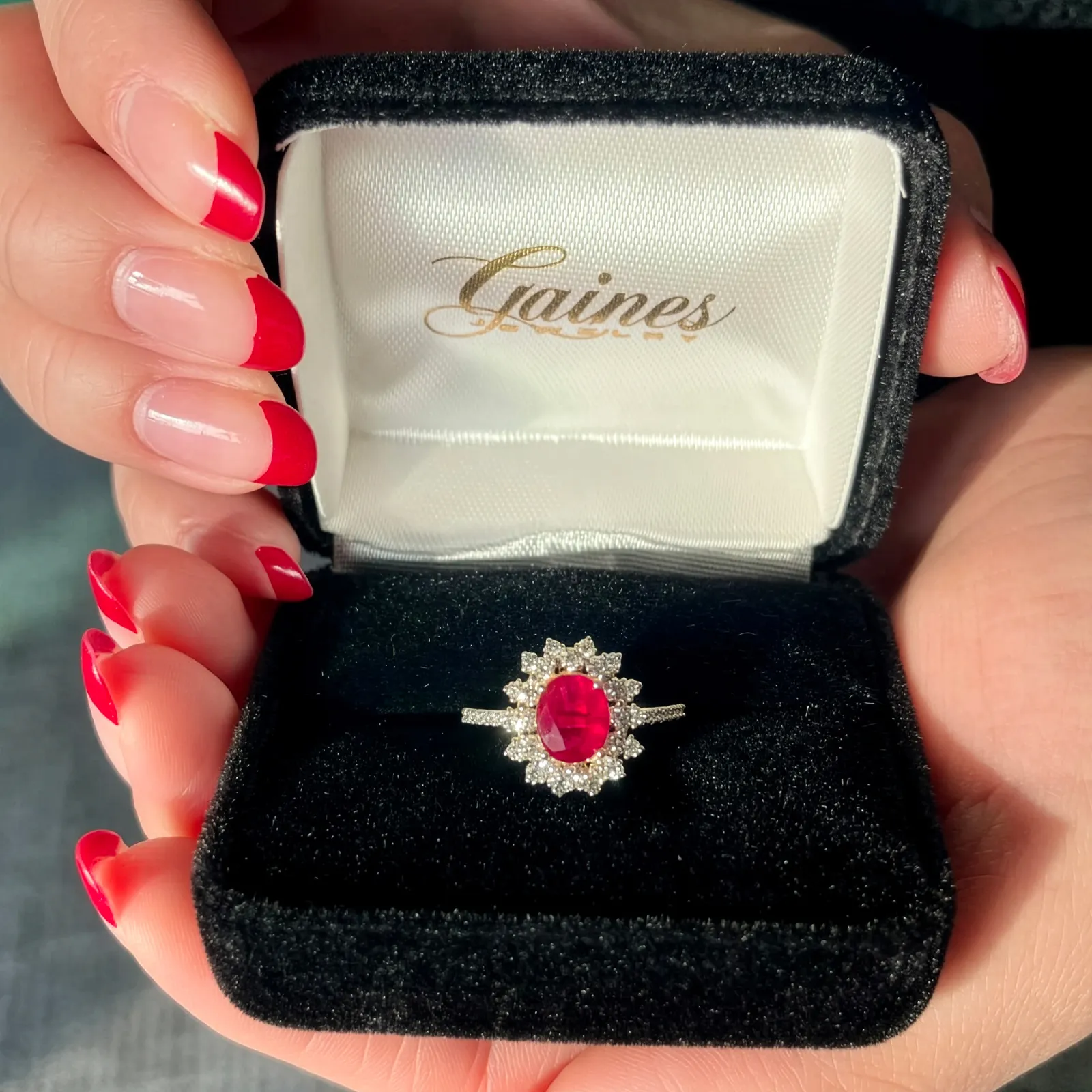 A hand with red nails holds a black box containing a ruby and diamond ring from Gaines Jewelry.