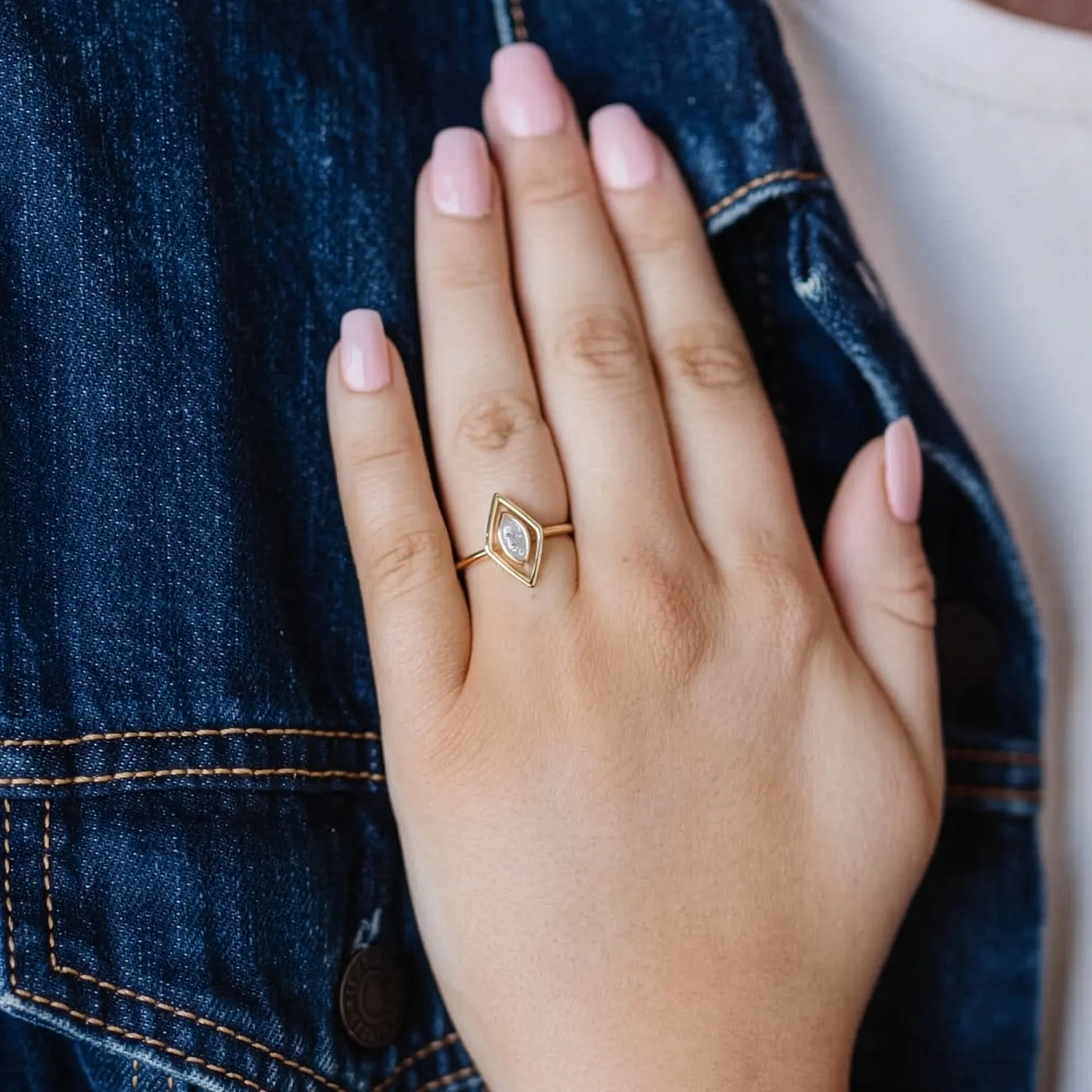 A hand with pink nails wearing a gold ring with a diamond, resting on a denim jacket.