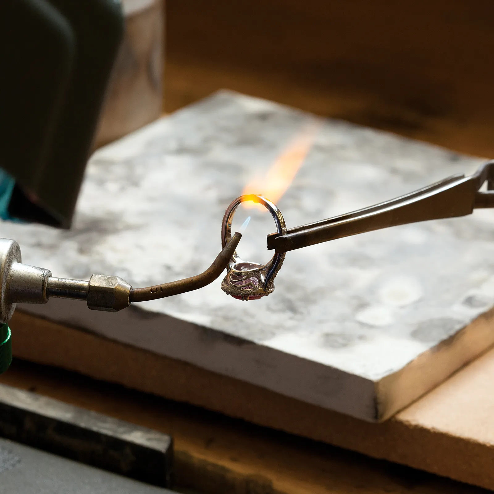 A jeweler uses a torch to solder a ring held by tweezers on a workbench.