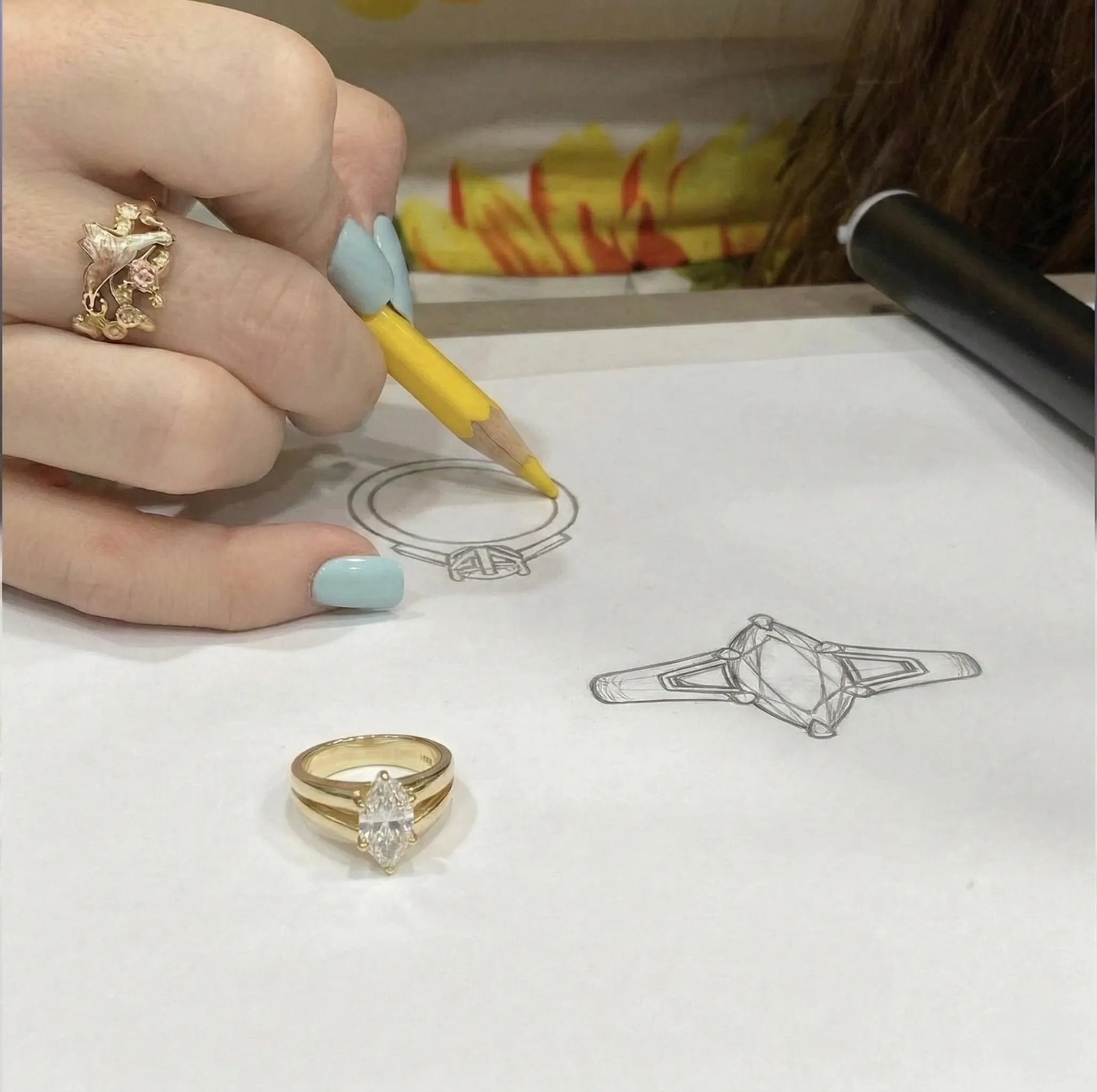 A hand sketches a ring design on paper with a pencil, next to a gold ring.