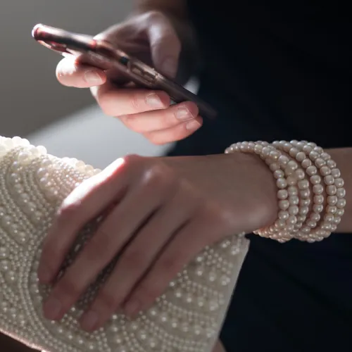 Lifestyle photo of a person holding a smartphone and a pearl-embellished clutch purse while wearing a layered multi-strand pearl wrap bracelet.