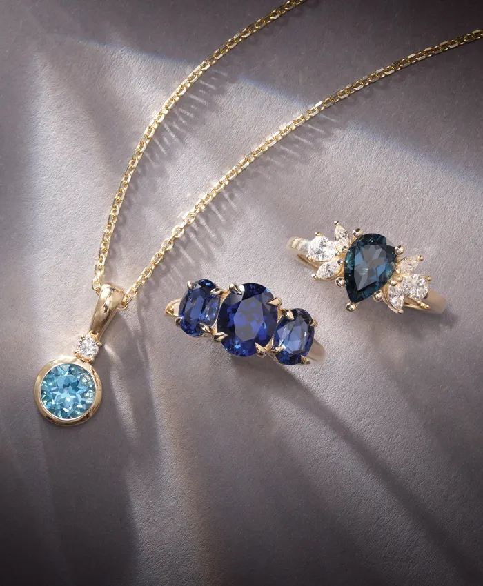 Elegant gold jewelry featuring a blue gemstone pendant and rings with sapphires and diamonds on a dark background.