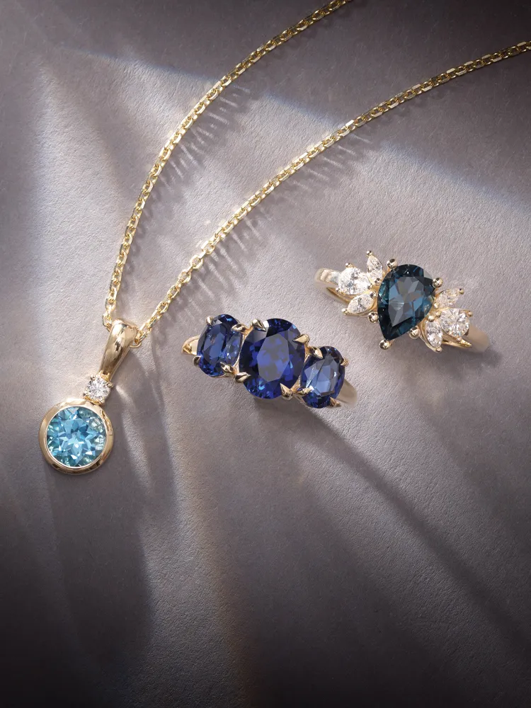 Elegant gold jewelry featuring a blue gemstone pendant and rings with sapphires and diamonds on a dark background.