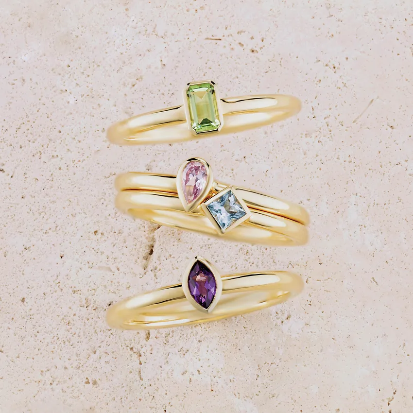 Three gold rings featuring colorful gemstones: green, pink, blue, and purple, displayed on a light textured background.