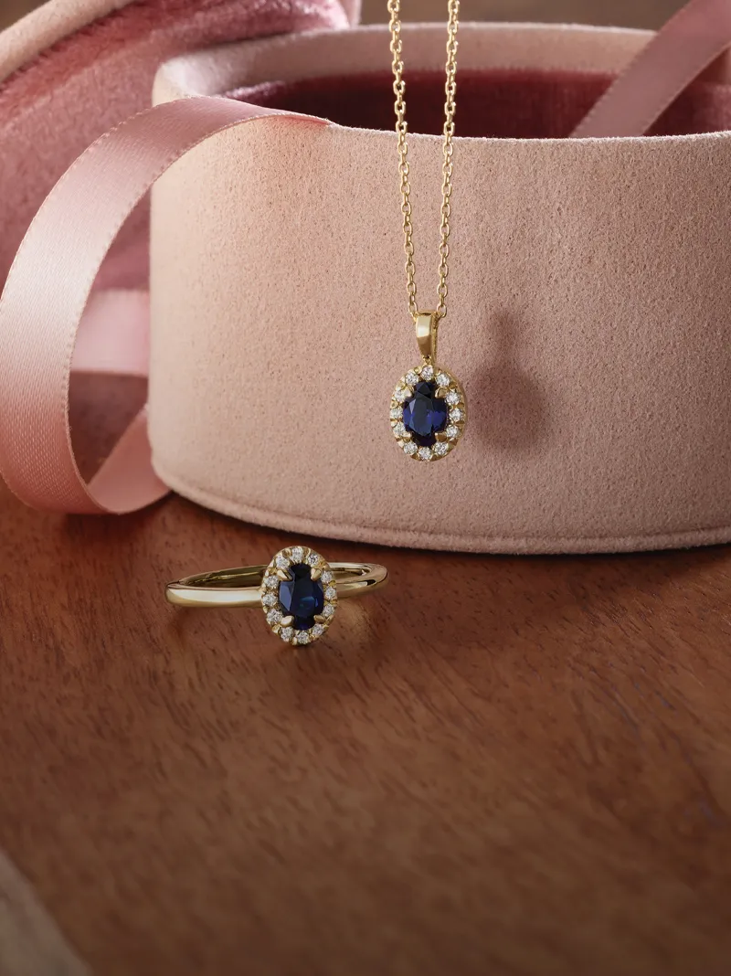 Gold sapphire pendant and ring set displayed on a wooden surface next to a pink jewelry box.