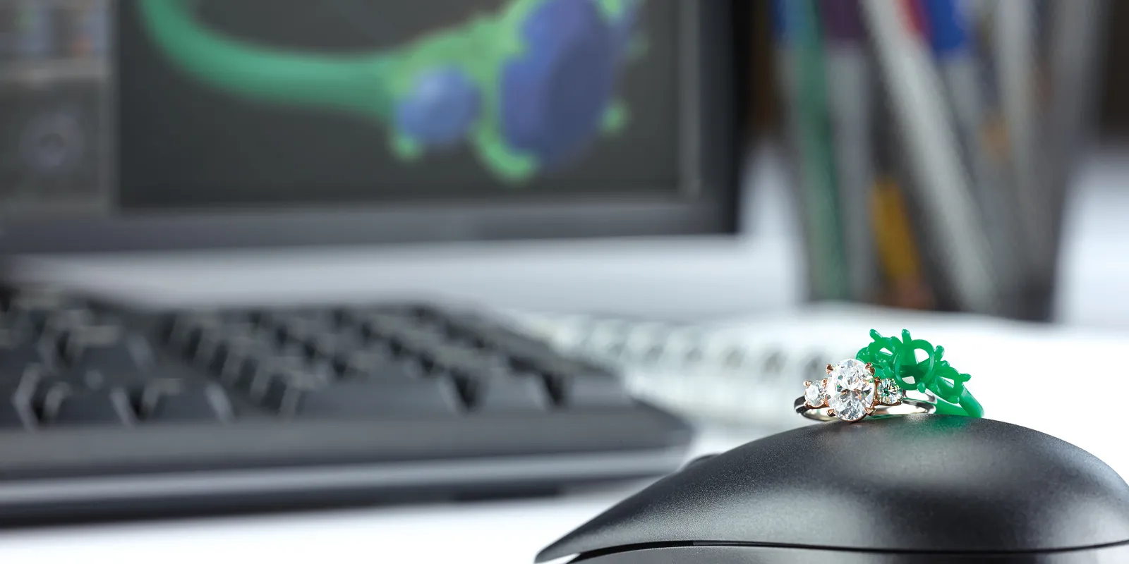 Engagement ring and green wax model on a computer mouse, with a design software screen in the background.
