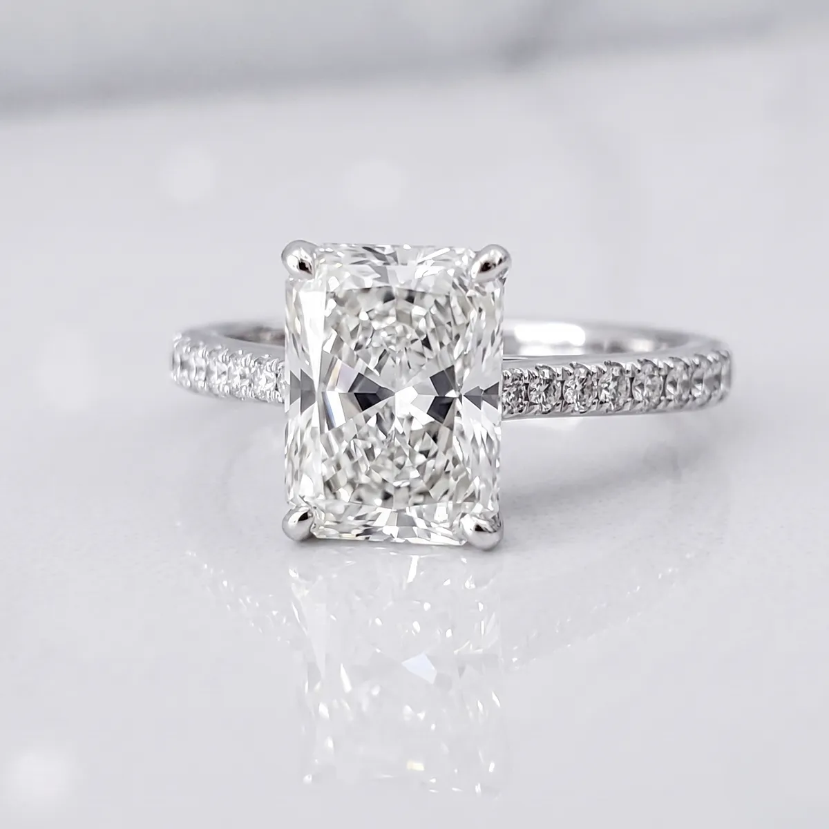 Radiant-cut diamond ring with a pavé band on a reflective surface.