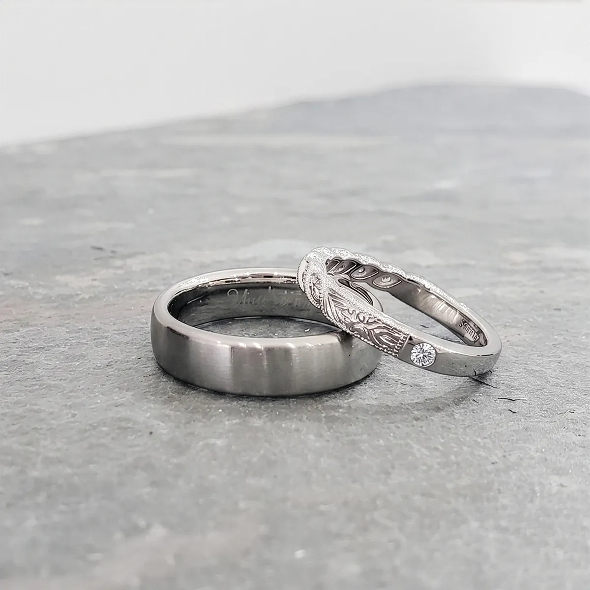 Two silver rings on a stone surface; one is plain, the other has intricate designs and a small diamond.