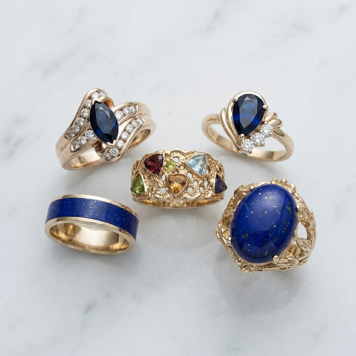 Five gold rings with blue and multicolored gemstones on a marble surface.