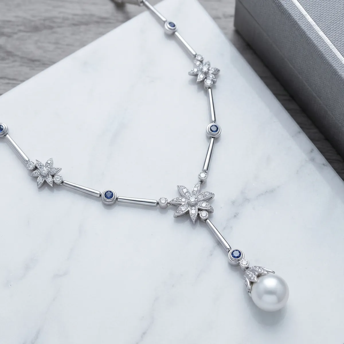 Elegant silver necklace with floral designs, blue stones, and a pearl pendant on a marble surface.