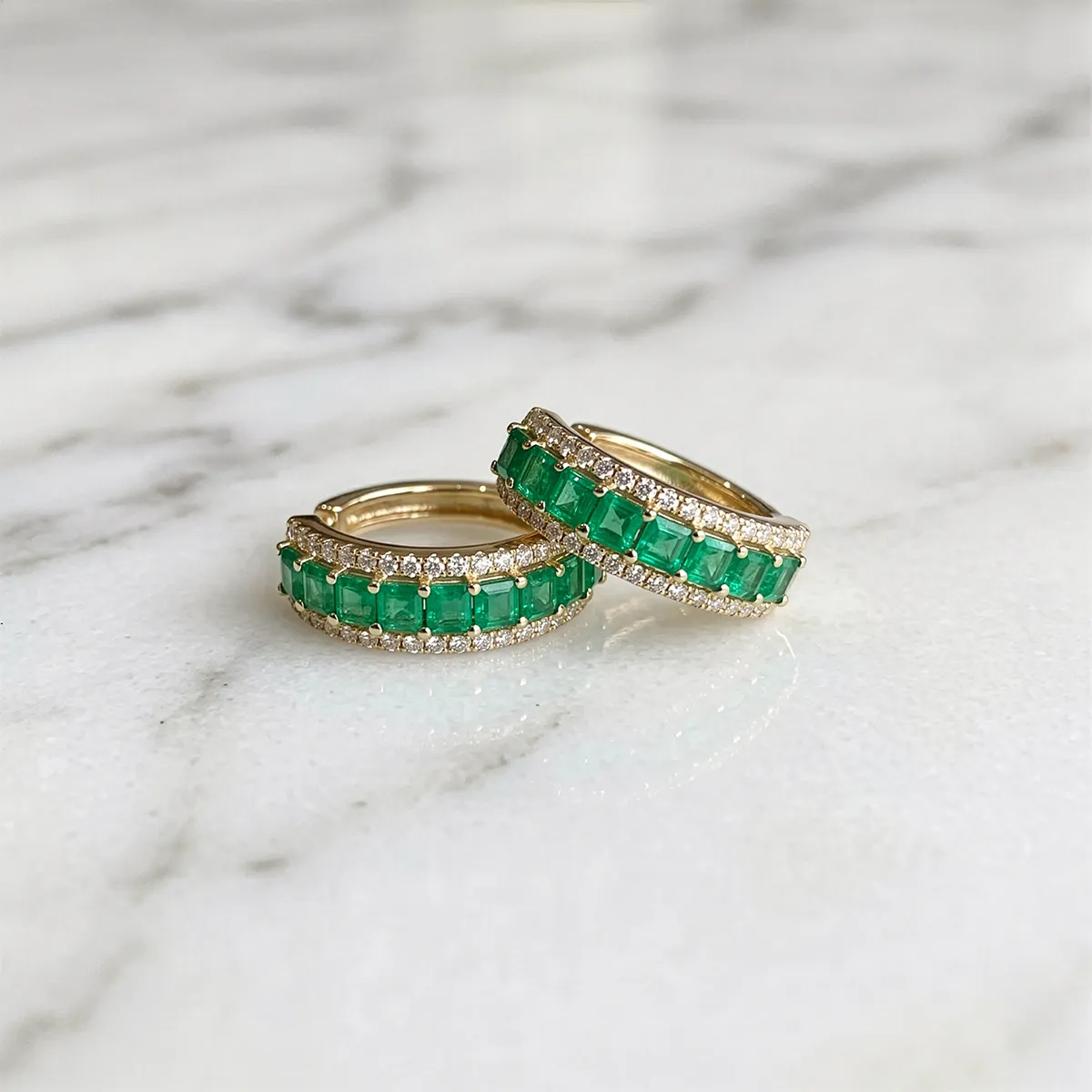 Emerald and diamond hoop earrings on a marble surface. Shop earrings at Georgetown Jewelers.