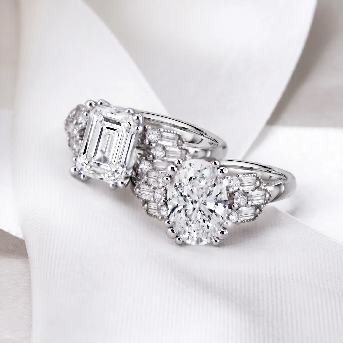 Two elegant diamond rings with emerald and oval cuts on a white fabric background. Shop engagement rings at Georgetown Jewelers.