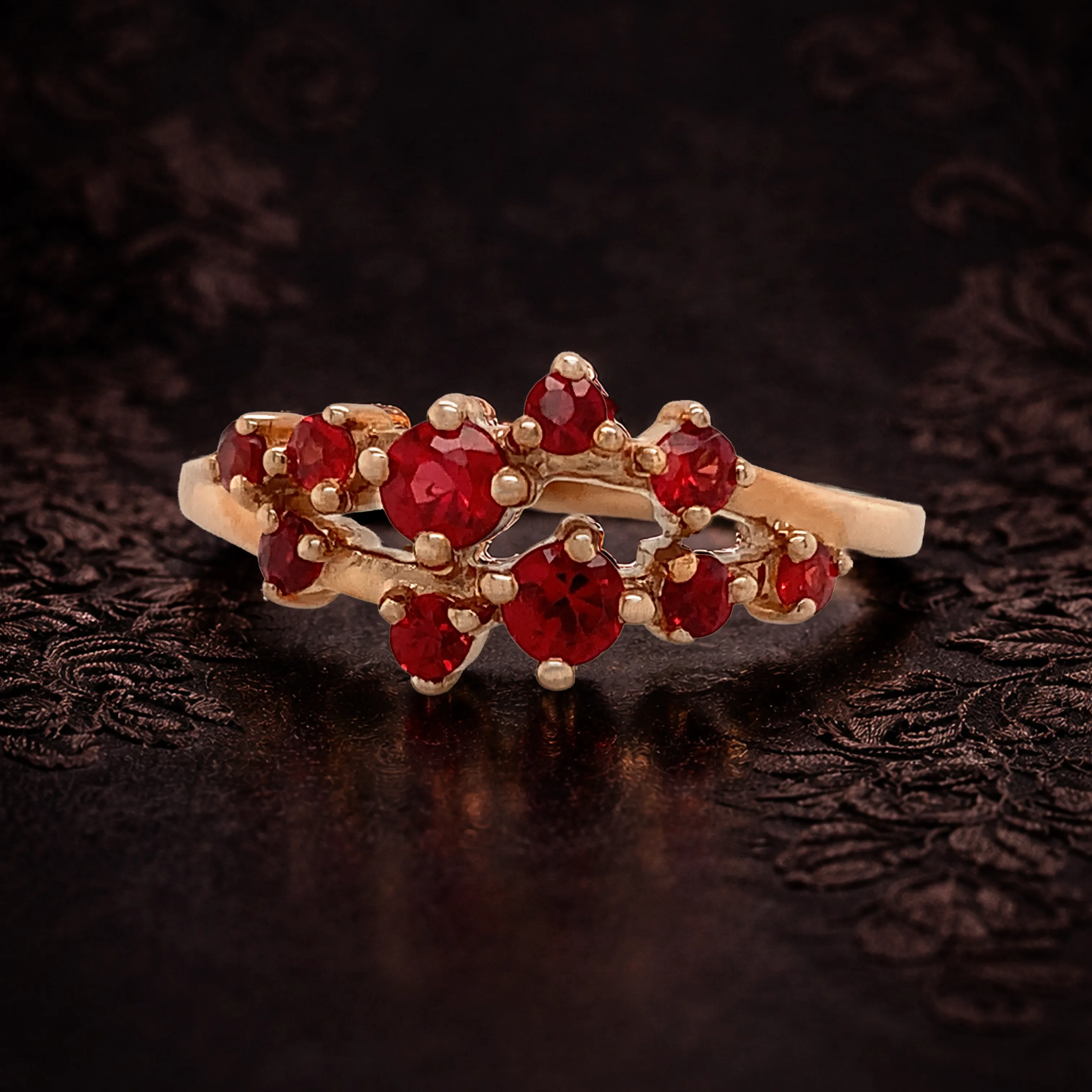 Gold ring with a red gemstone and diamond accents on a dark floral background.