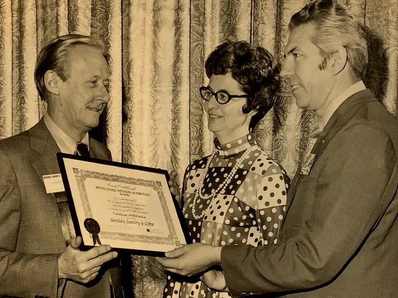Three people in formal attire holding a framed certificate in front of curtains.