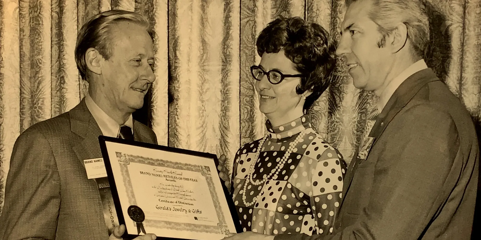 Three people in formal attire holding a framed certificate in front of curtains.