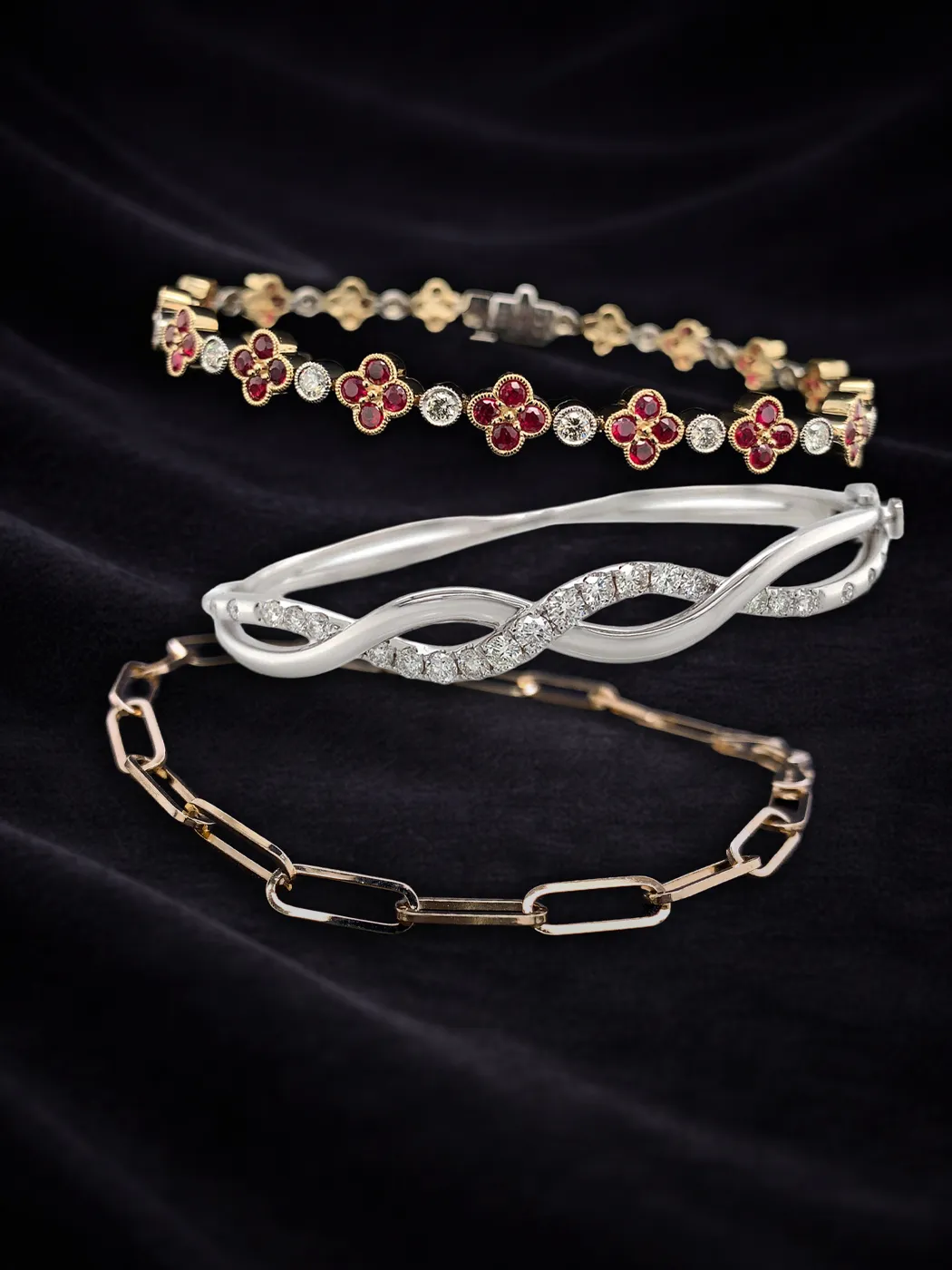 Three elegant bracelets on black fabric: floral, diamond twist, and chain link designs.