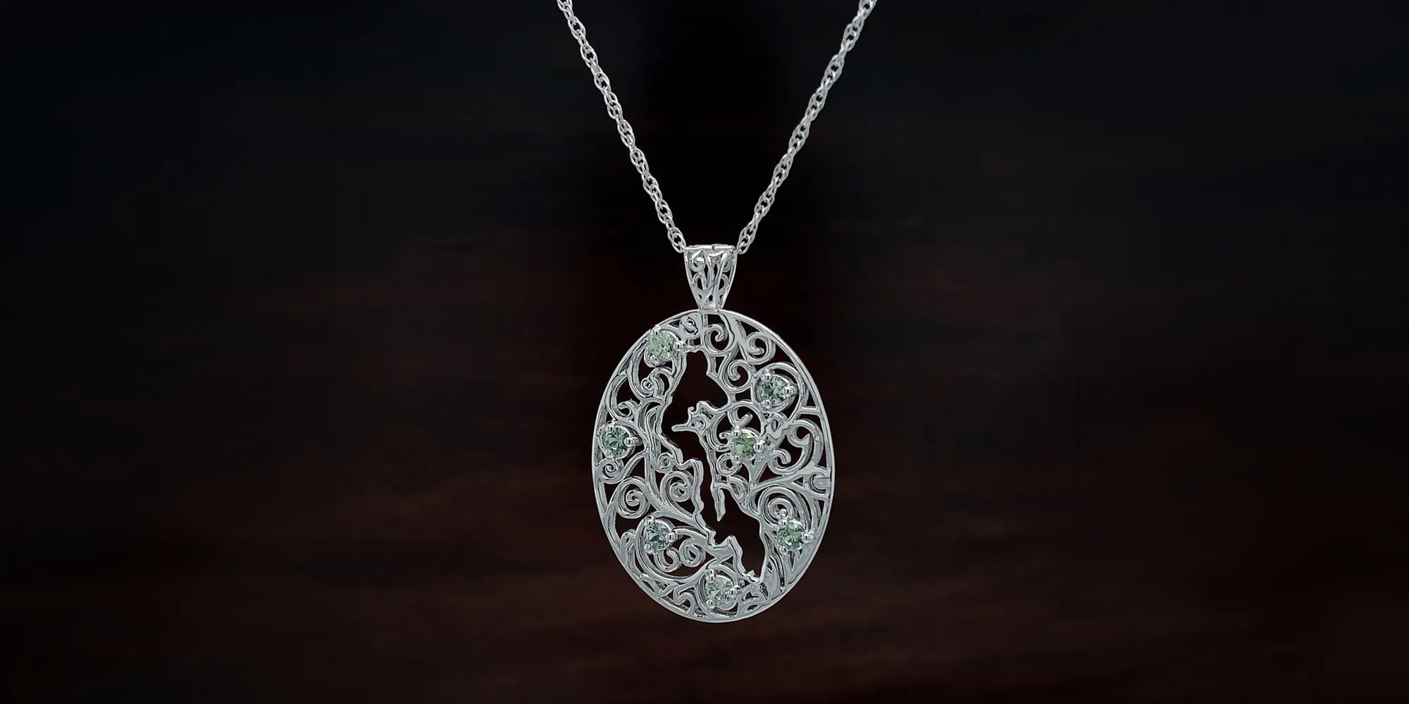 Ornate silver pendant with intricate swirls and green gemstones on a chain, set against a dark background.