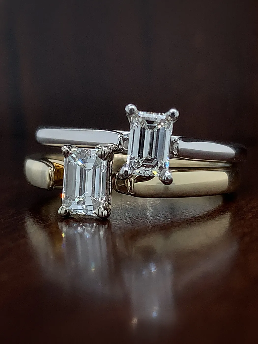 Two engagement rings with emerald-cut diamonds on a dark surface.