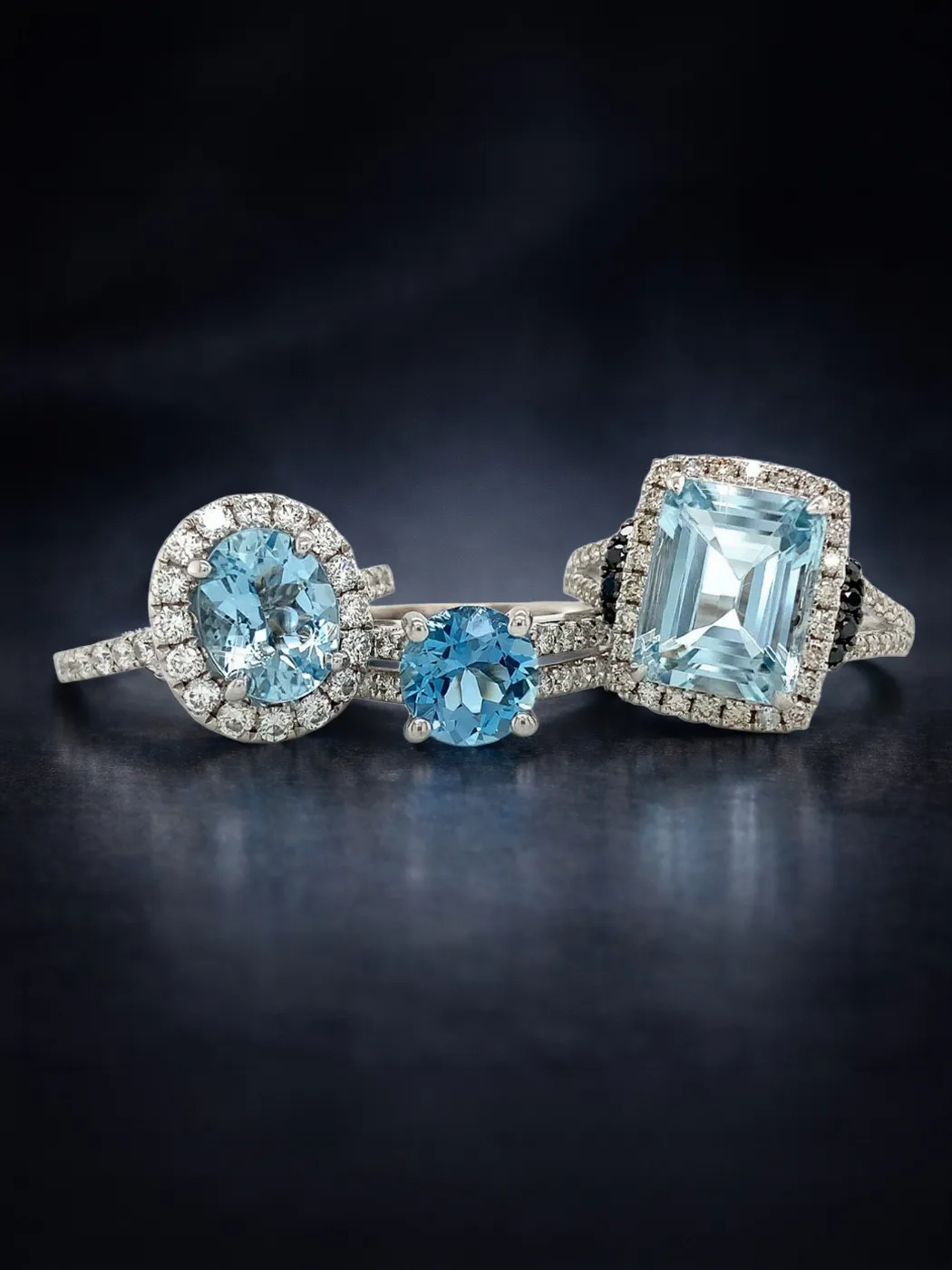 Three aquamarine rings with diamond accents on a dark background.