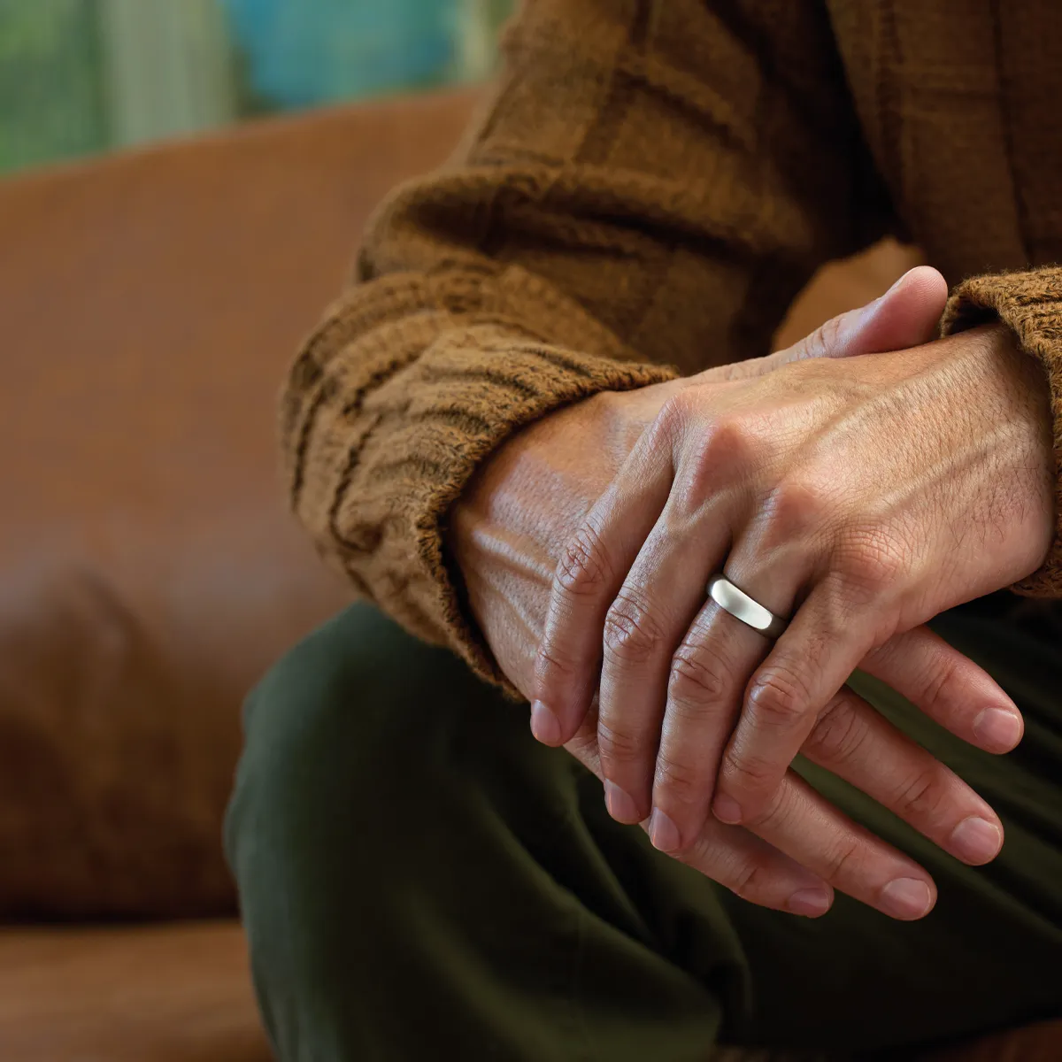 Close-up of a person's hands with a silver ring, resting on a brown sweater, against a soft-focus background.