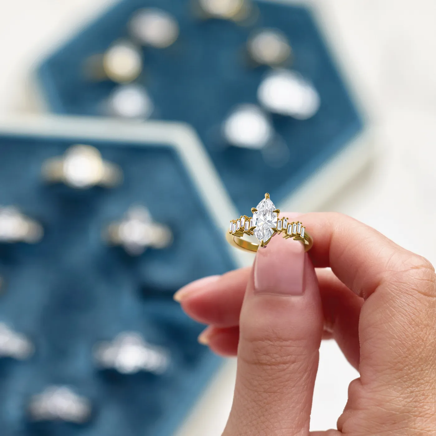 Hand holding a gold engagement ring with a marquise diamond, surrounded by other rings on a blue velvet display.
