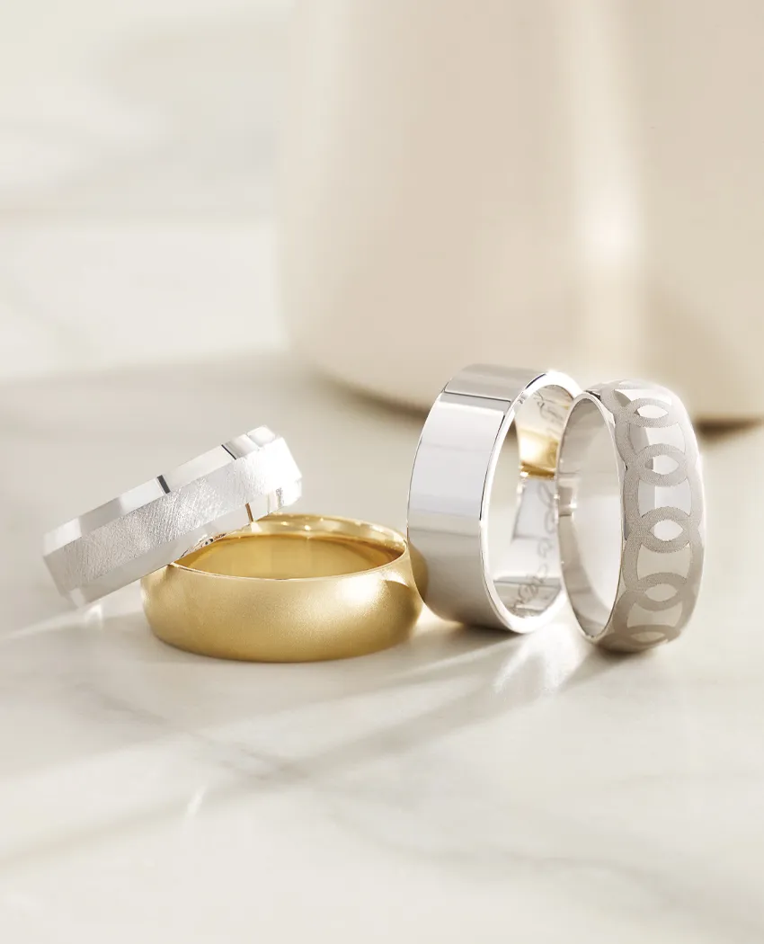 Four stylish wedding bands in gold and silver tones displayed on a marble surface. Click to explore Wedding Bands at Golden Gift