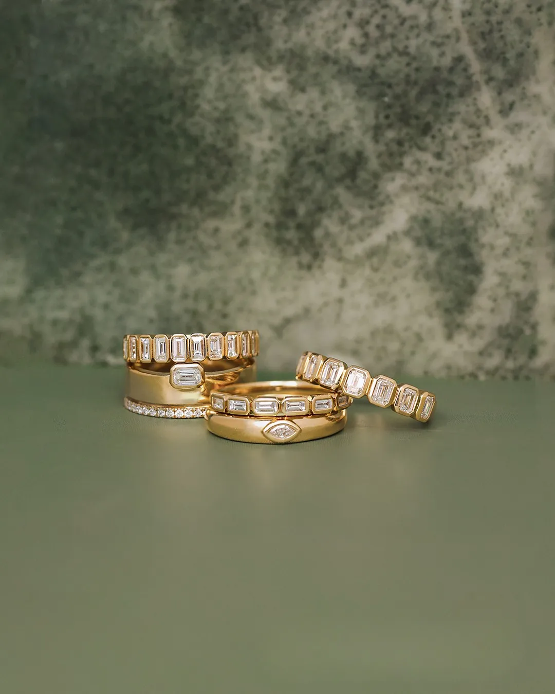 Four elegant gold rings with various designs displayed on a green background. Shop fine rings at Gray's Jewelers Bespoke.