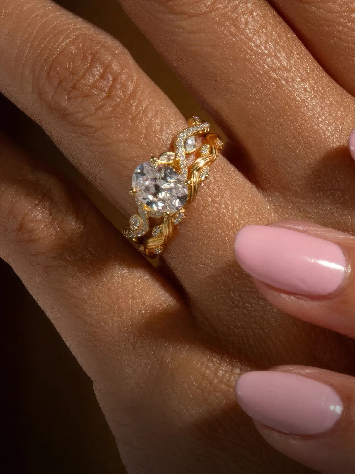A hand with pink nails wearing a gold ring with a large diamond.
