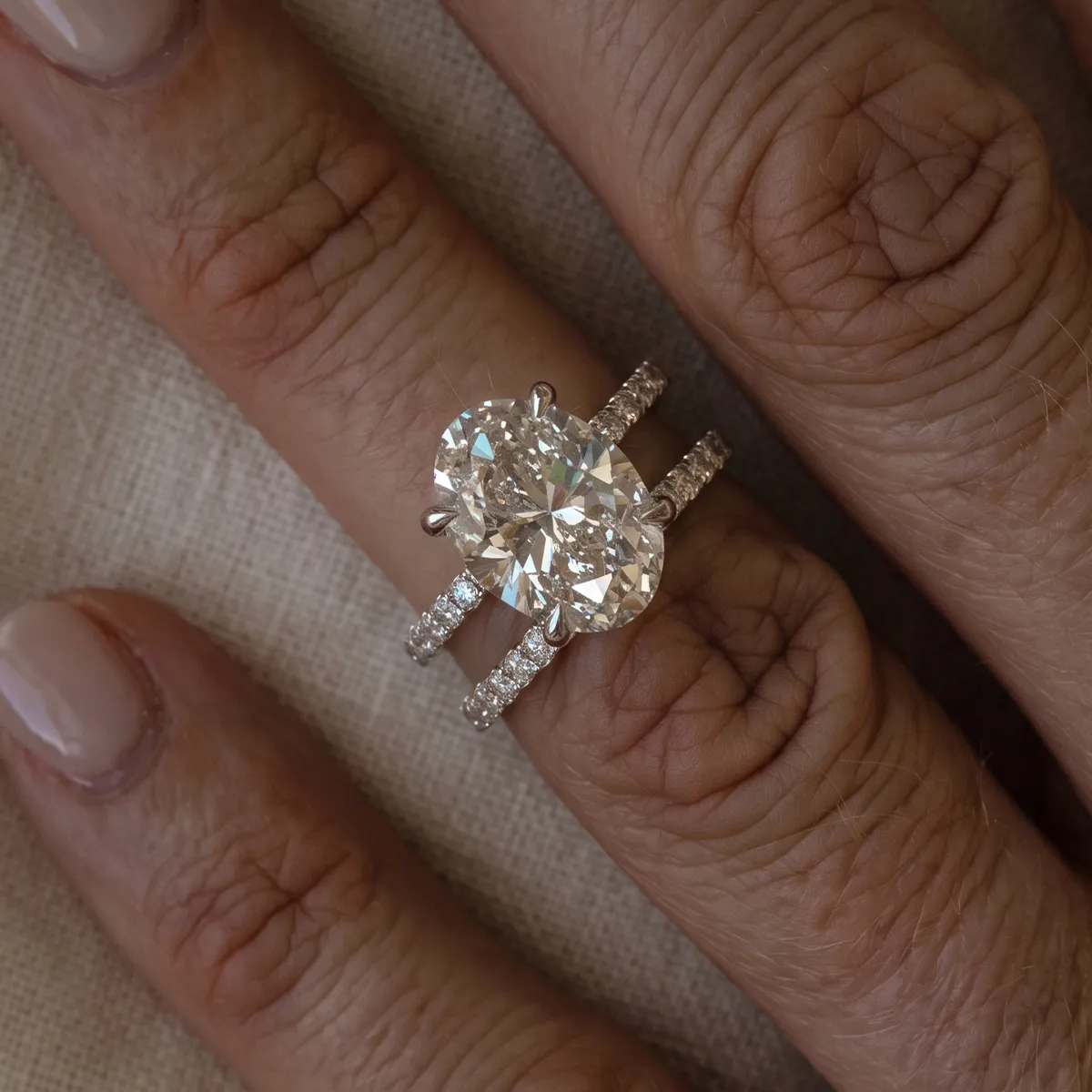 A hand wearing a large oval diamond ring with a matching diamond band on a textured background.