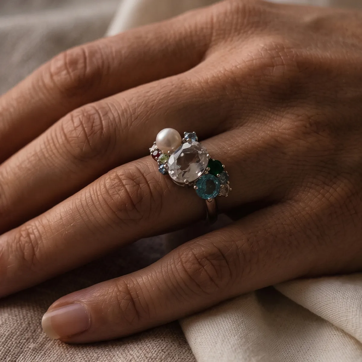 A hand wearing a ring with a pearl and various gemstones on a textured fabric background.