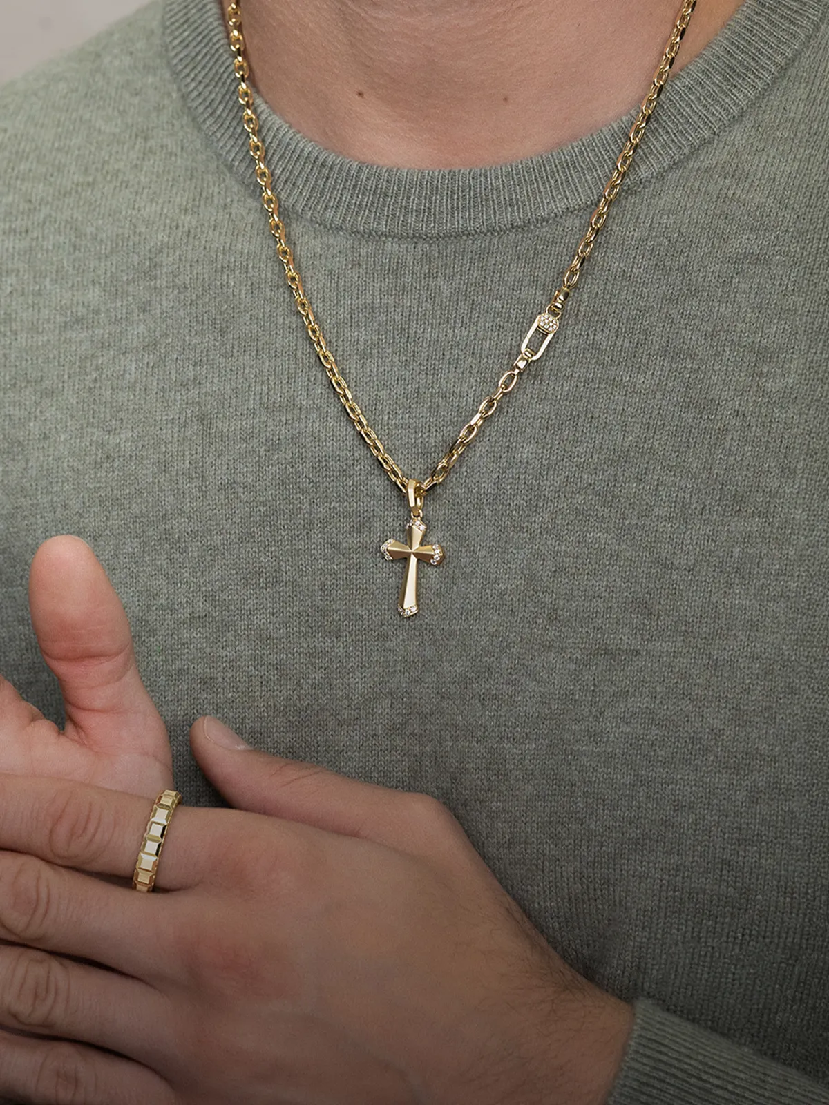 A person wearing a gold chain with a cross pendant and a gold ring, dressed in a gray sweater.
