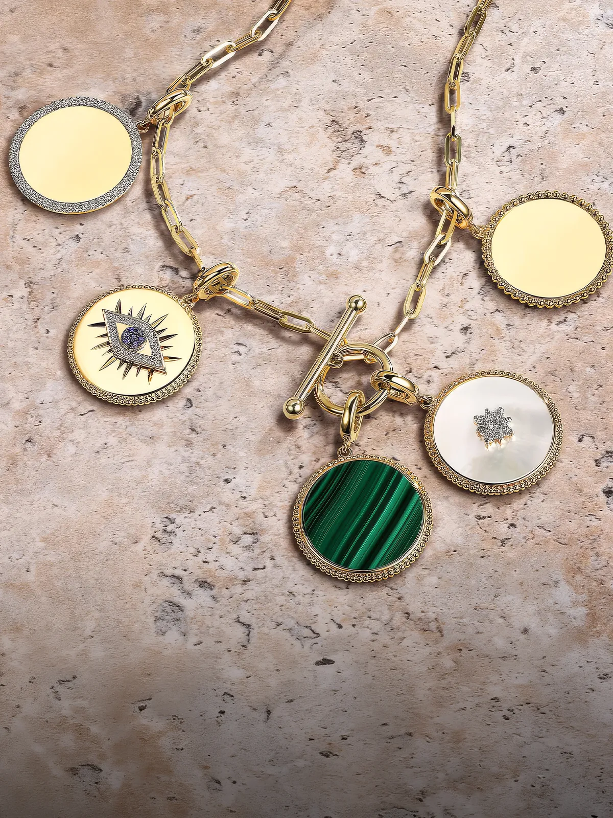 Gold chain necklace with five round charms, including a green stone and an eye design, on a textured surface.