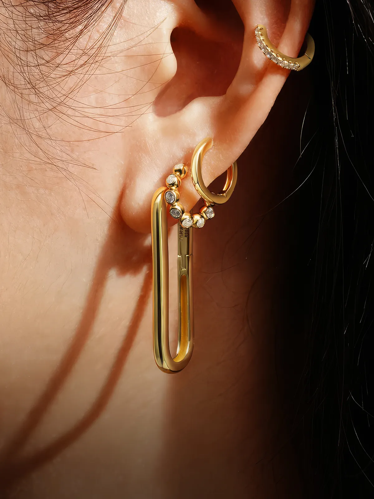 Close-up of an ear with multiple gold and diamond earrings, including a large paperclip-style earring.