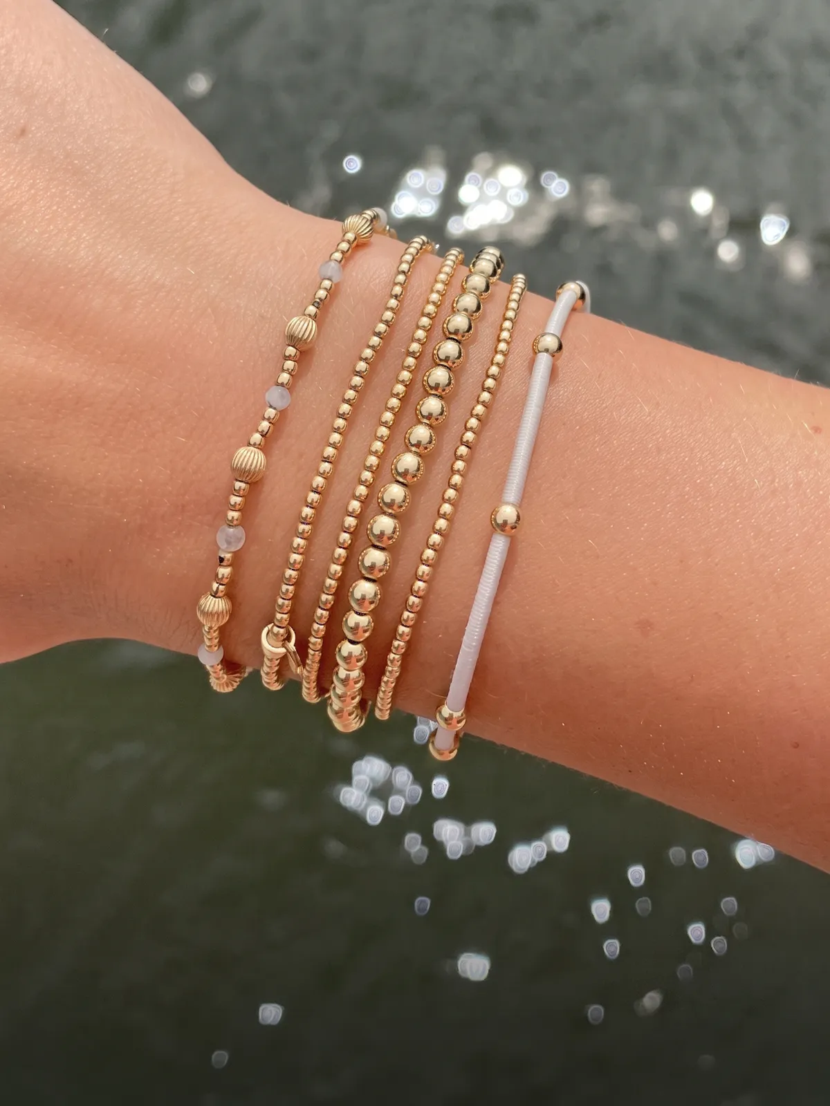 A wrist adorned with multiple gold and pearl bracelets against a blurred water background.