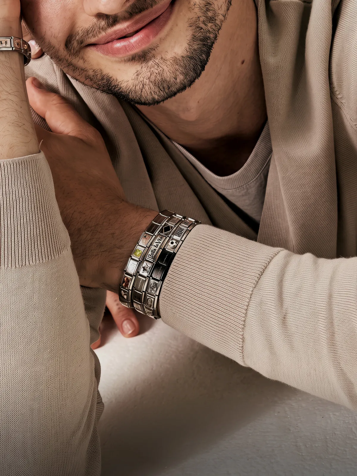 A man wearing multiple silver bracelets and a beige sweater, smiling slightly.