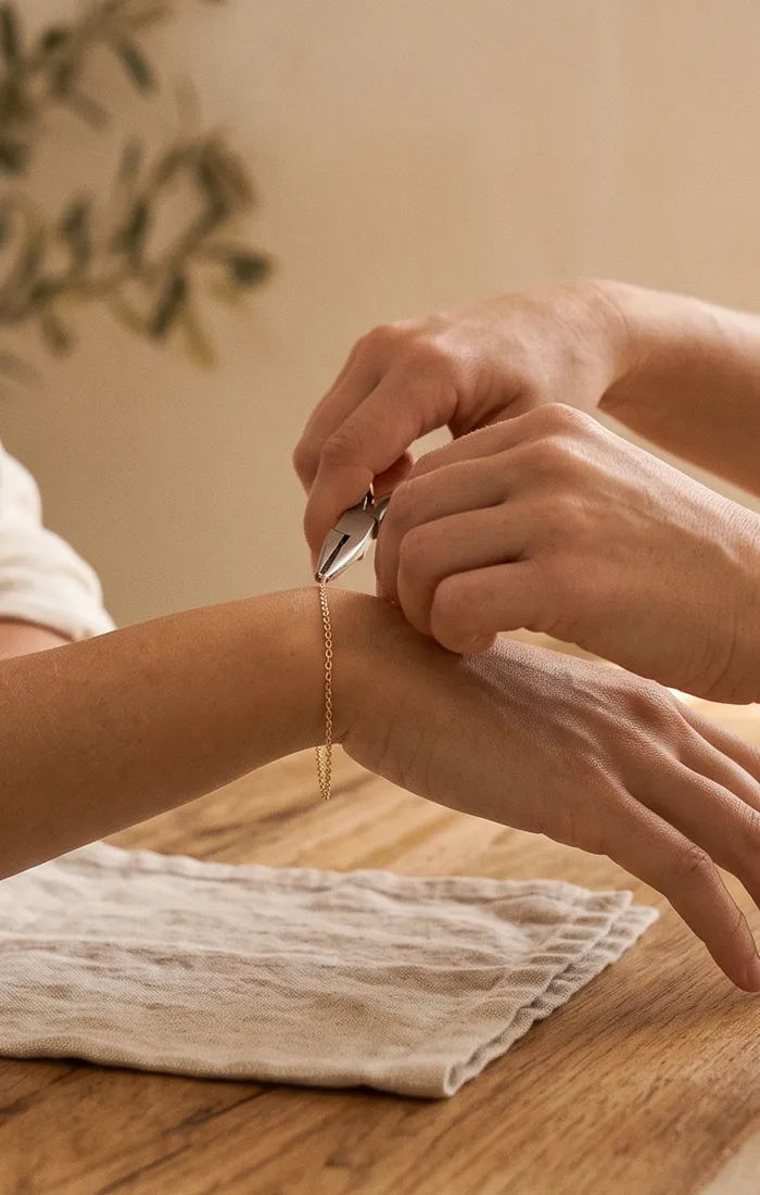 Two hands working on a delicate gold chain with pliers on a wooden table.