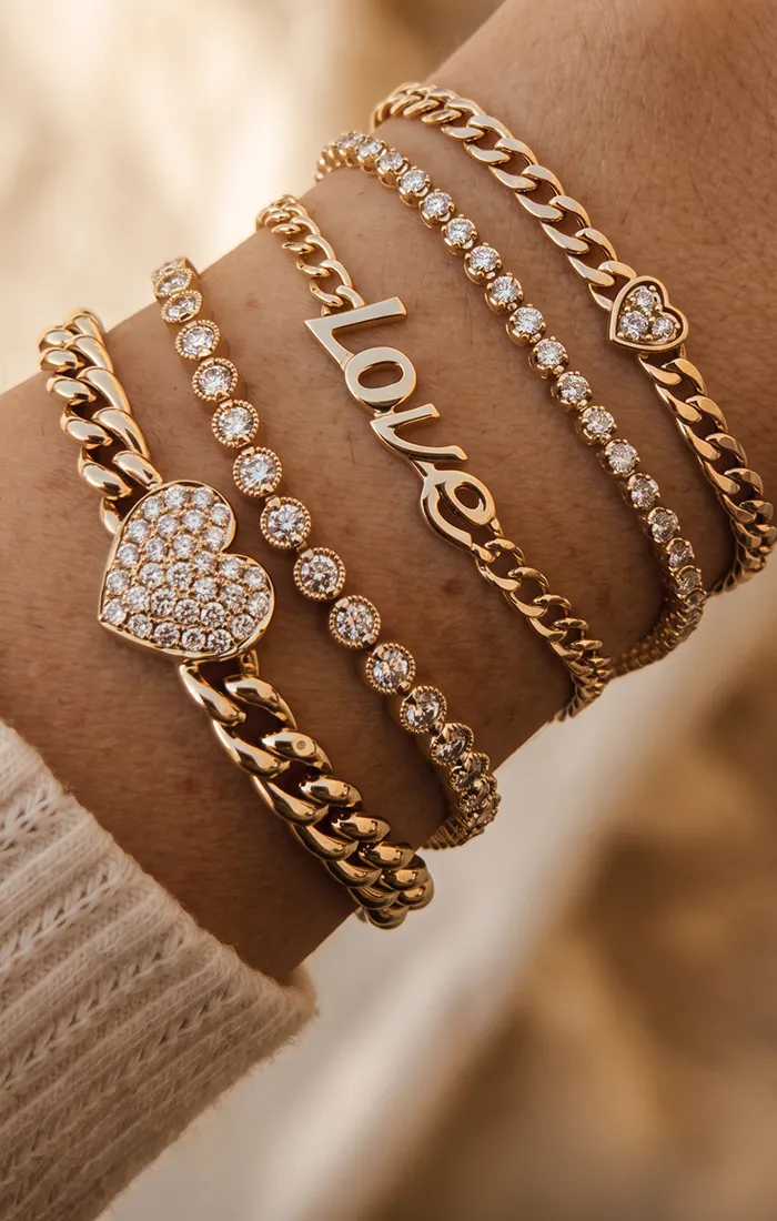 A wrist adorned with multiple gold bracelets featuring hearts, diamonds, and the word 